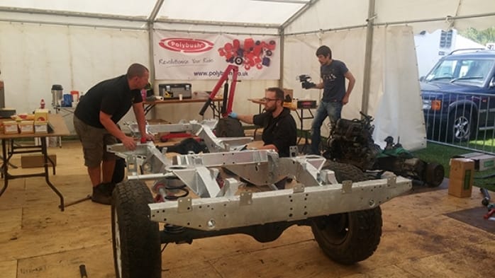 Three men work on a car chassis in a workshop marquee with various car parts and a camera setup; a banner with "polybush" hangs in the background.