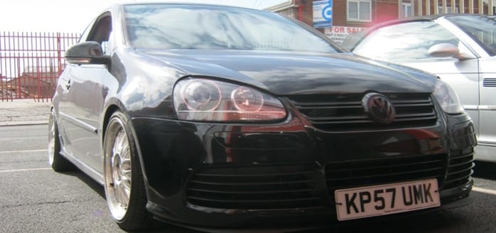 A low-angle view of a black Volkswagen car, parked on a street. The car features a distinctive front grille and round headlights, with a visible number plate reading "kp57 umk".