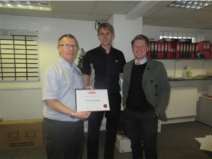 Three blokes in an office environment, grinning as the taller chap in the middle brandishes a certificate. In the backdrop, there are shelves filled with files.