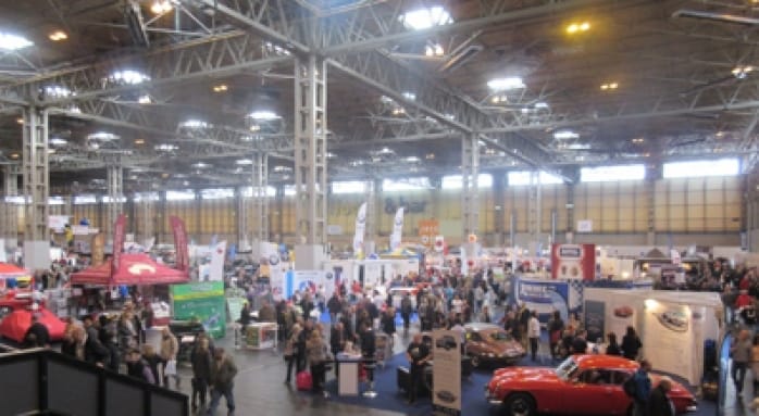 A busy indoor exhibition with various stalls, displays, and a throng of visitors. There are cars on show, promotional banners, and different trade stands in a large hall with high ceilings.