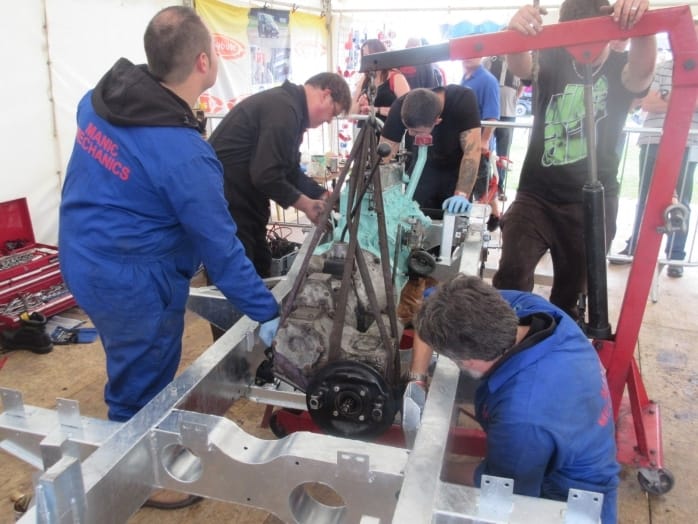 Five mechanics collaborate to fit an engine into a vehicle chassis in a workshop marquee, encapsulated by tools and parts.
