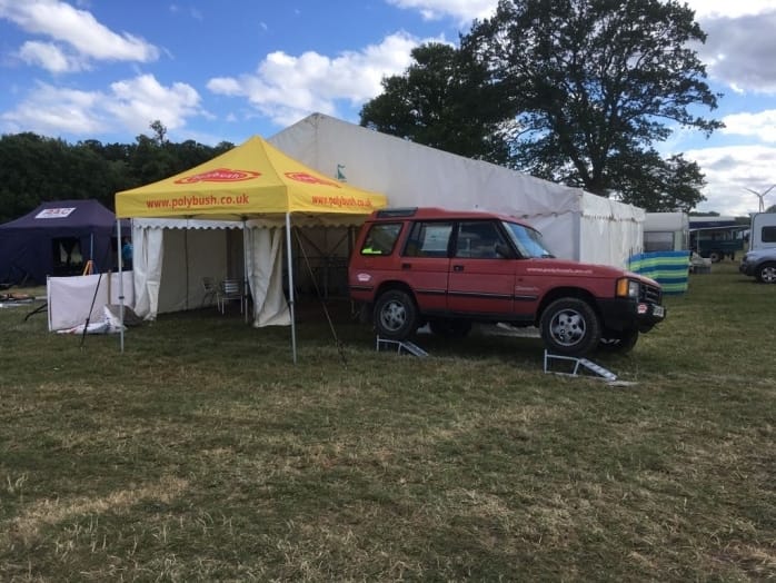 A red land rover parked beside a commercial marquee with banners at an outdoor event, with other marquees and vehicles in the background under a cloudy sky.