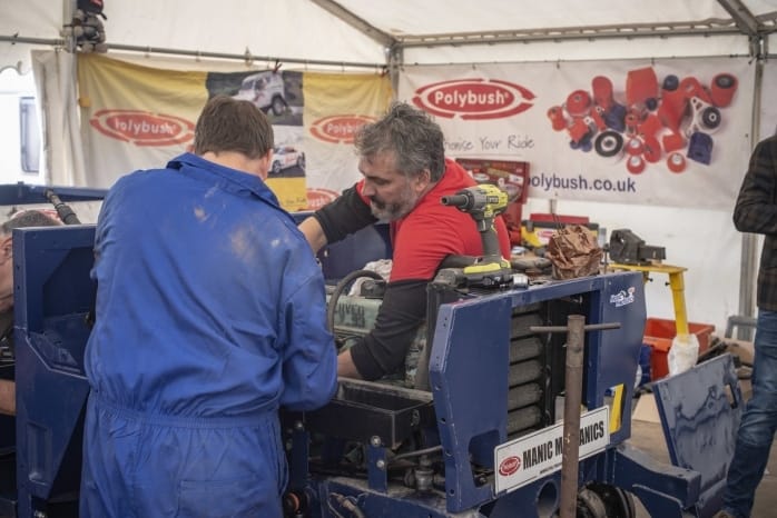Two mechanics in blue coveralls working on a car engine in a marquee at an outdoor event, with a banner for "polybush" in the background.