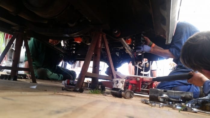 Three mechanics work beneath a hoisted car, utilising tools to mend or inspect its underside. The emphasis is on their hands and apparatus, with particulars of the vehicle's suspension visible.