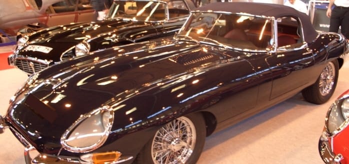 Two shiny classic Jaguar E-type cars displayed at a car exhibition, featuring sleek black and red models with chrome detailing and wire wheels.