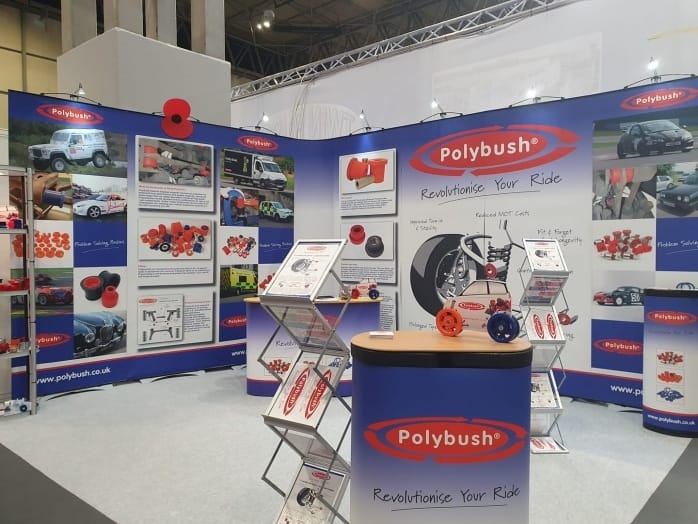 A exhibition stand for polybush featuring displays with car parts, informational panels, and promotional materials. A lectern in the front displays various auto components.
