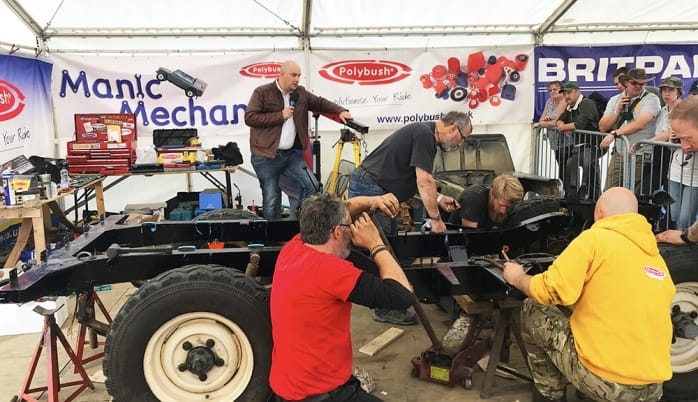 A group of individuals are observing a mechanic work on a vehicle chassis at an outdoor event beneath a marquee with various car parts and sponsor banners displayed in the backdrop.