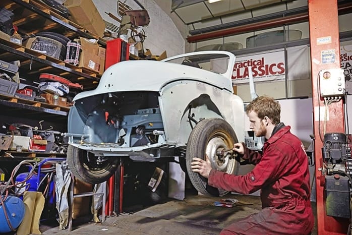 A mechanic in a red boiler suit works on restoring a classic white car in a cluttered workshop filled with shelves of parts and tools.