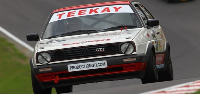 A vintage Volkswagen Golf GTI race car speeds on a racecourse, prominently featuring sponsor logos "teekay" and "productiongti.com" on a grey day.