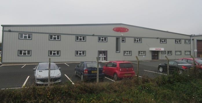 A large industrial grey building with a red logo on top, a car park in front with various cars, and some greenery in the foreground on a foggy day.