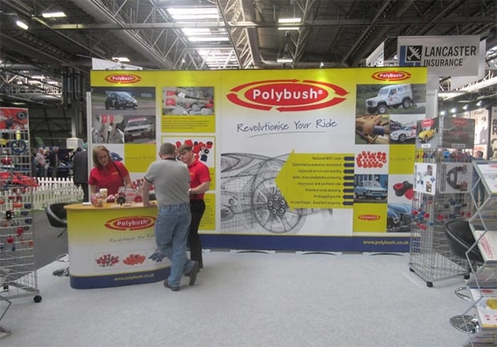 Two people stand at the polybush exhibition booth with car part images and text, at an automotive trade fair. A banner with the slogan "revolutionise your ride" is displayed prominently.
