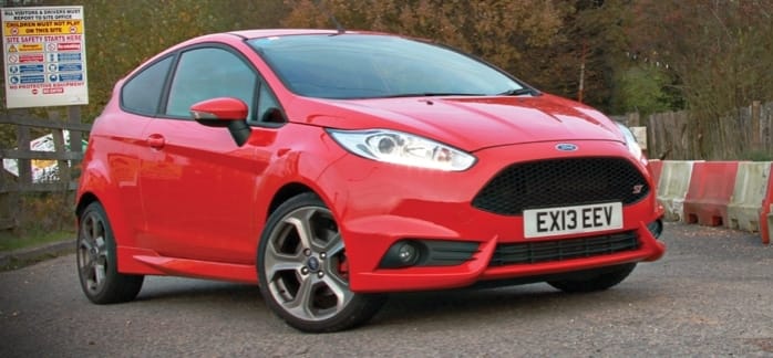 A bright red Ford Fiesta parked outside, boasting a sleek design with stylish alloy wheels, situated in front of a fence with parking notices.