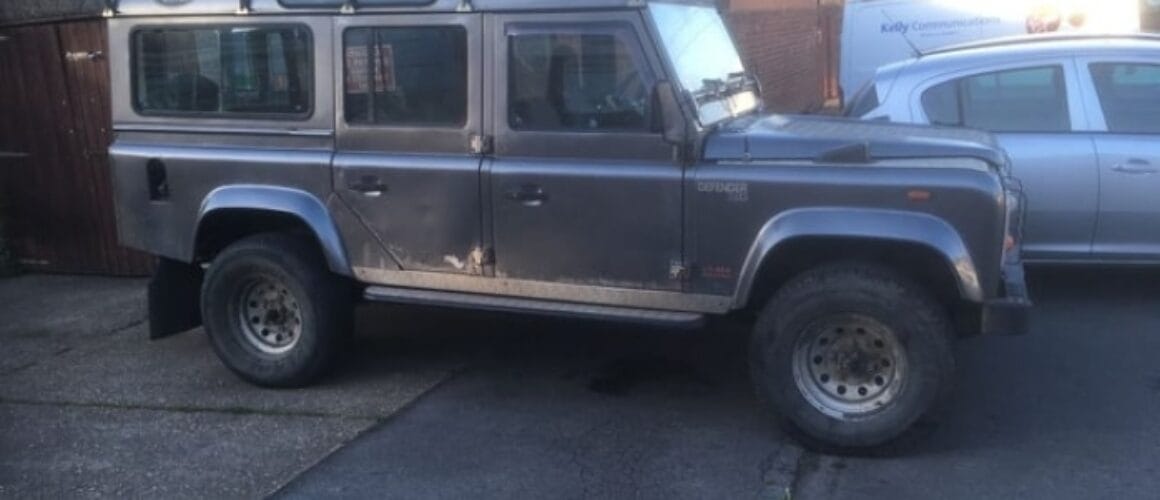 A grey land rover defender parked diagonally on a narrow street, with a wooden garage and another car in the background.