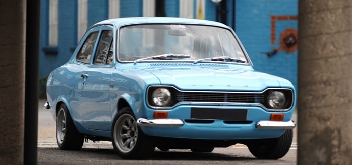 A classic blue Ford Escort car parked on a street, observed between two pillars, in an urban setting with brick walls and a hint of graffiti.