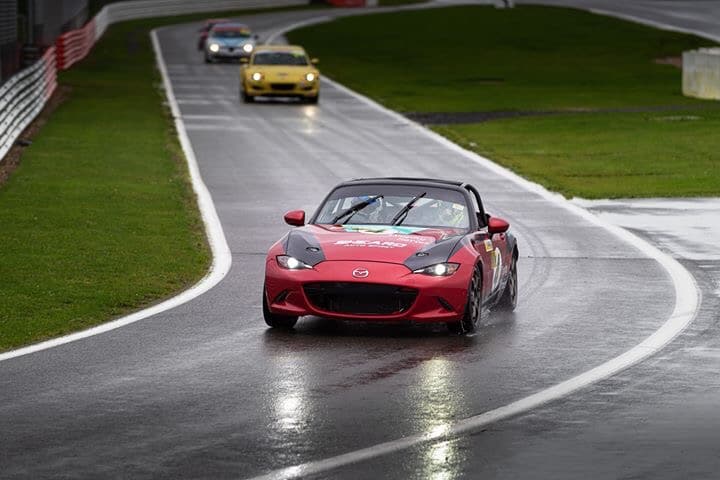 A red Mazda convertible leads a race on a damp circuit, followed by several other sports cars, including a yellow one, at a motorsport event.