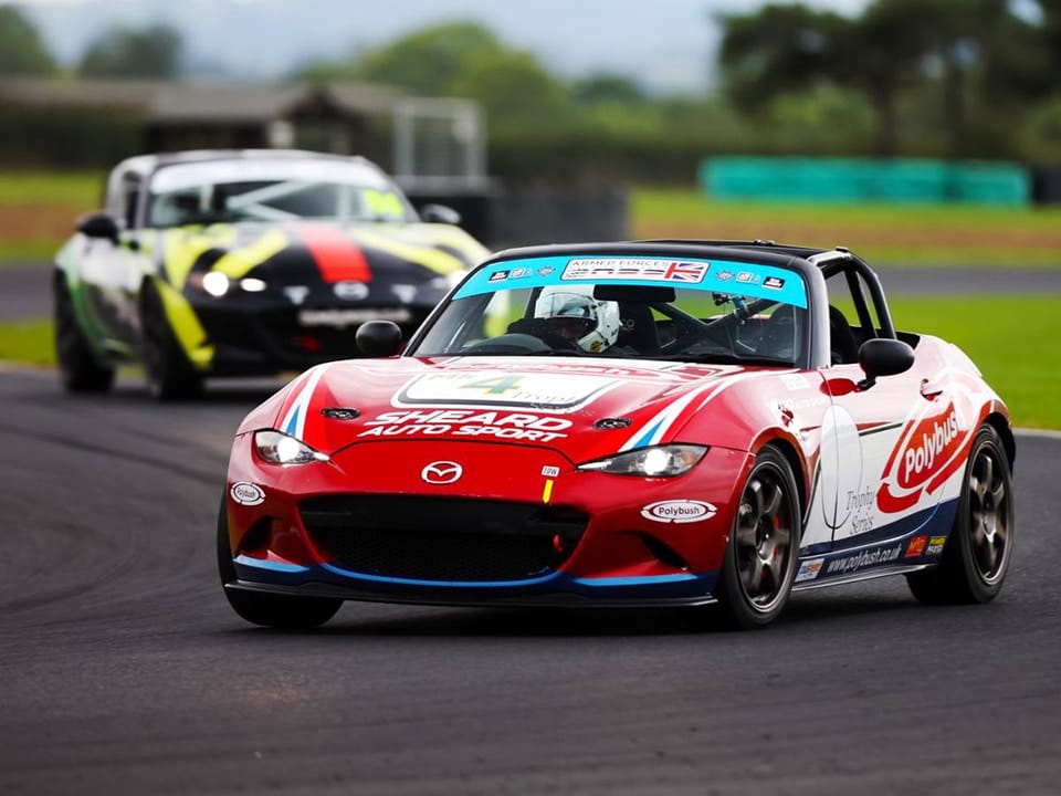 A red racing car with sponsor logos leads another car on a tarmac racetrack, showcasing dynamic motion and competitive racing.