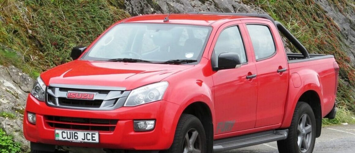 A red Isuzu D-Max pick-up truck parked on a road near a rocky hillside, featuring a clean exterior and visible number plate "cu16 jce".
