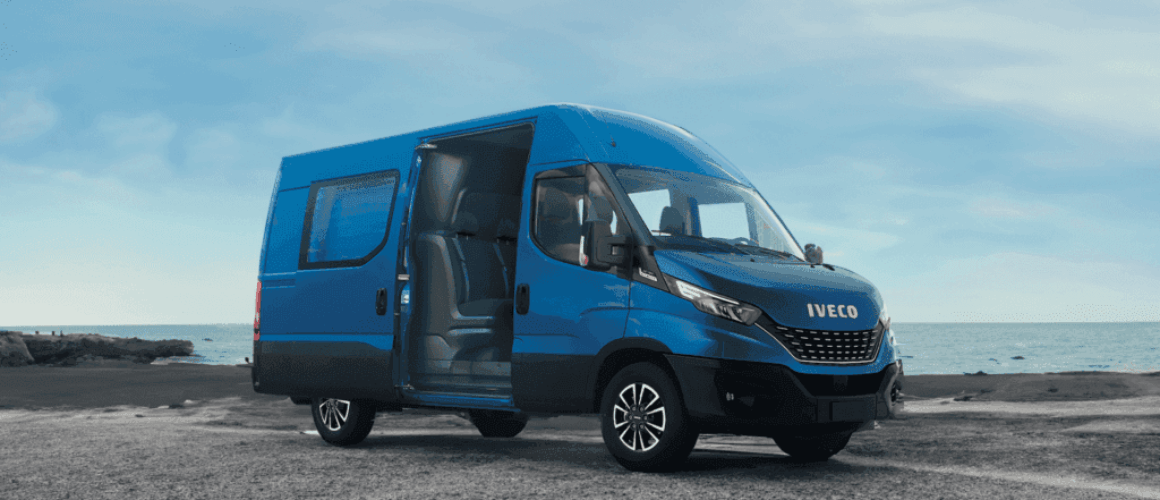 A blue Iveco van parked by the seaside with the driver's door open, showing the interior. The backdrop features a clear sky and rocky coastline.
