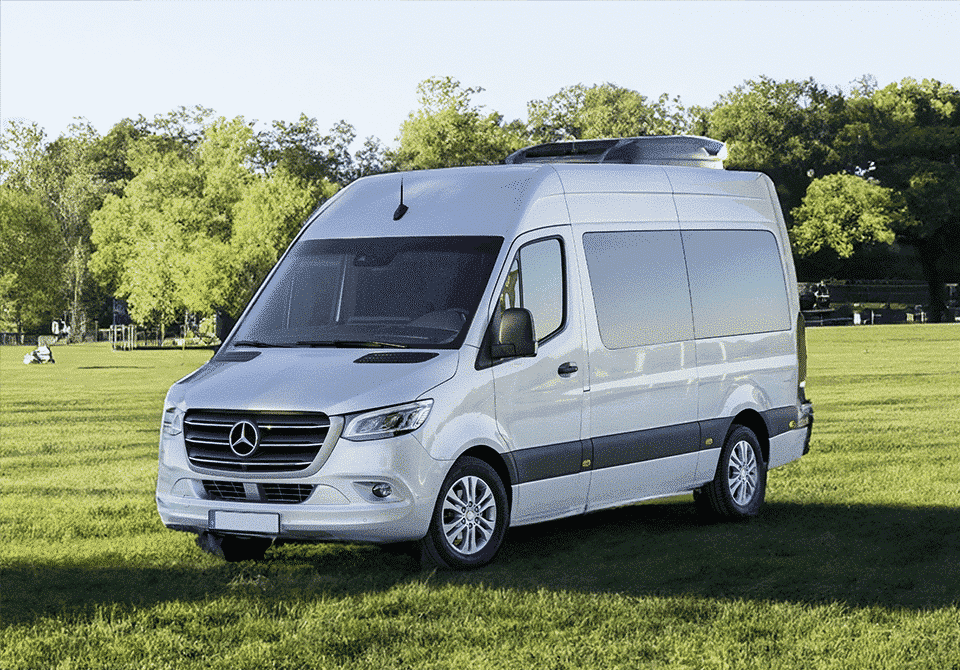A white Mercedes van parked on a grassy field in a park, with trees and a person sitting in the background under the shade.(This sentence does not contain any American English-specific terms or spellings that need to be translated into British English.)