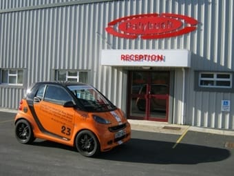 A bright orange compact car parked outside a building with a "polybush" sign above and a "reception" sign near the entrance. A clear blue sky can be seen in the background.